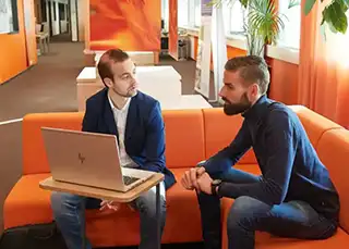 Twee mannen met elkaar in gesprek op een oranje bank achter een laptop.