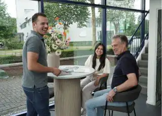 Drie mensen rondom een tafel die met elkaar lachen en vrolijk zijn.